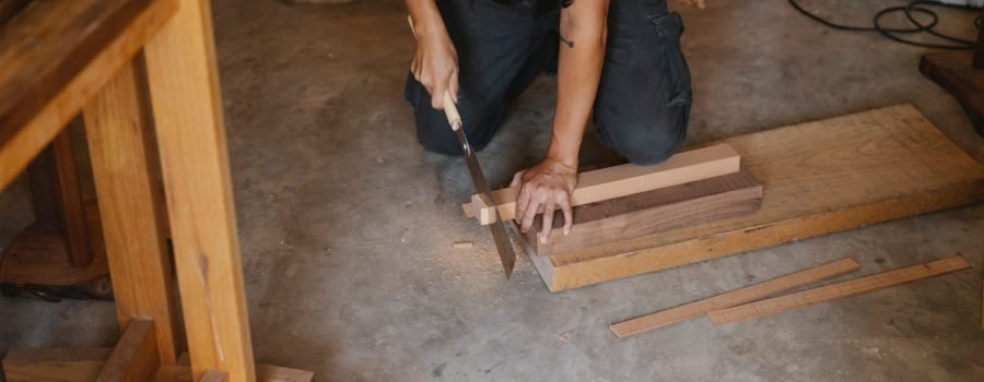 Anonymous joiner working with Japanese saw while cutting edge of timber plank on floor in workshop